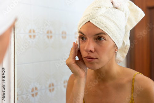 una mujer entre 20 o 30 años haciéndose limpieza de cutis frente al espejo con crema antienvejecimiento e hidratante. En un primer plano de su cara con 1 toalla en el pelo . photo