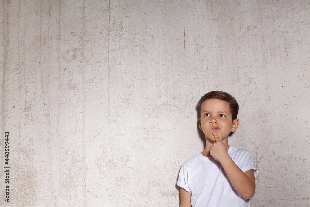 Pensive little guy stands on the wall background. Portrait of wistful boy in white T-shirt, copy space. Concept of reflections and looking for new idea. Pensive little guy stands on the wall background. Portrait of wistful boy in white T-shirt, copy space. Concept of reflections and looking for new idea.