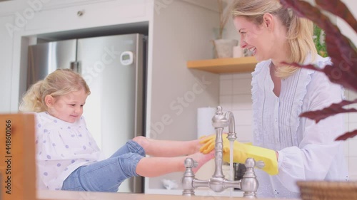 Mother wearing rubber gloves at home in kitchen with young daughter having fun as girl puts feet in sink as they do washing up at sink- shot in slow motion
