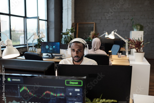 Young African businessman in headphones working in front of computer monitor in office photo