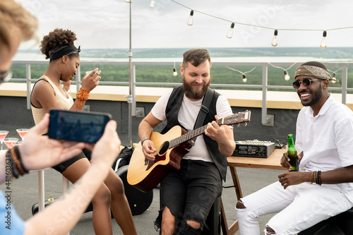 Multracial friends with drinks enjoying rooftop party while one of them shooting video