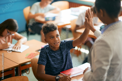 African American schoolboy and his teacher giving high five in classroom.