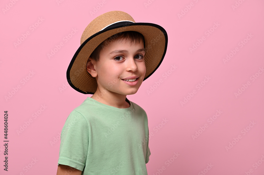 Side portrait of a handsome schoolboy in summer straw hat, looking at camera and standing isolated over pink background with copy space. Close-up Side portrait of a handsome schoolboy in summer straw hat, looking at camera and standing isolated over pink background with copy space. Close-up