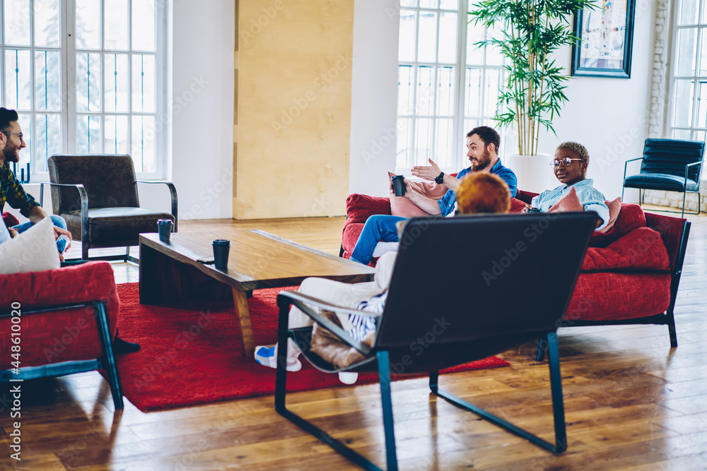 Group of multiracial people talking with friends sitting on comfortable red sofa enjoying coffee break in stylish home interior, young trendy people communicating in loft hostel apartment