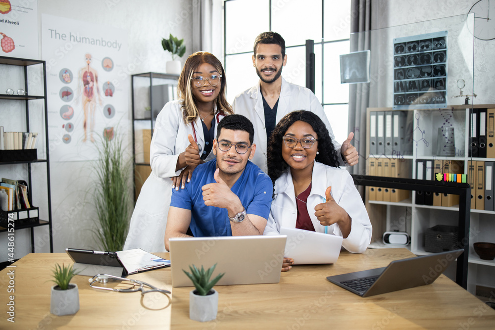 Fototapeta premium Multiracial group of doctors wearing coats and uniforms, sitting together at table and smiling on camera with thumbs up. Medical specialists during session at hospital. Cooperation concept.