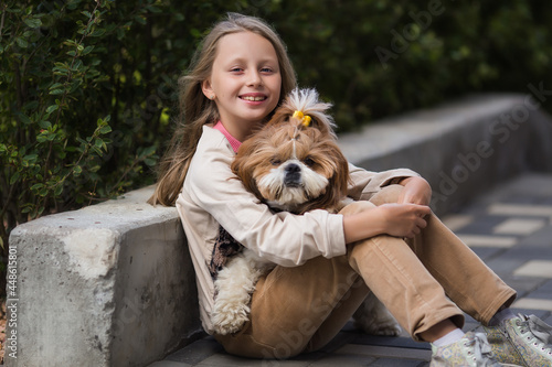 Portrait of beautiful preteen girl petting and hugging shih tzu dog looking at the camera outdoors.