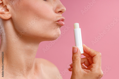 Close up shot of woman holding lip balm near her lips and making kissing gesture  posing isolated over pink background. Beauty  lip care concept. Side view