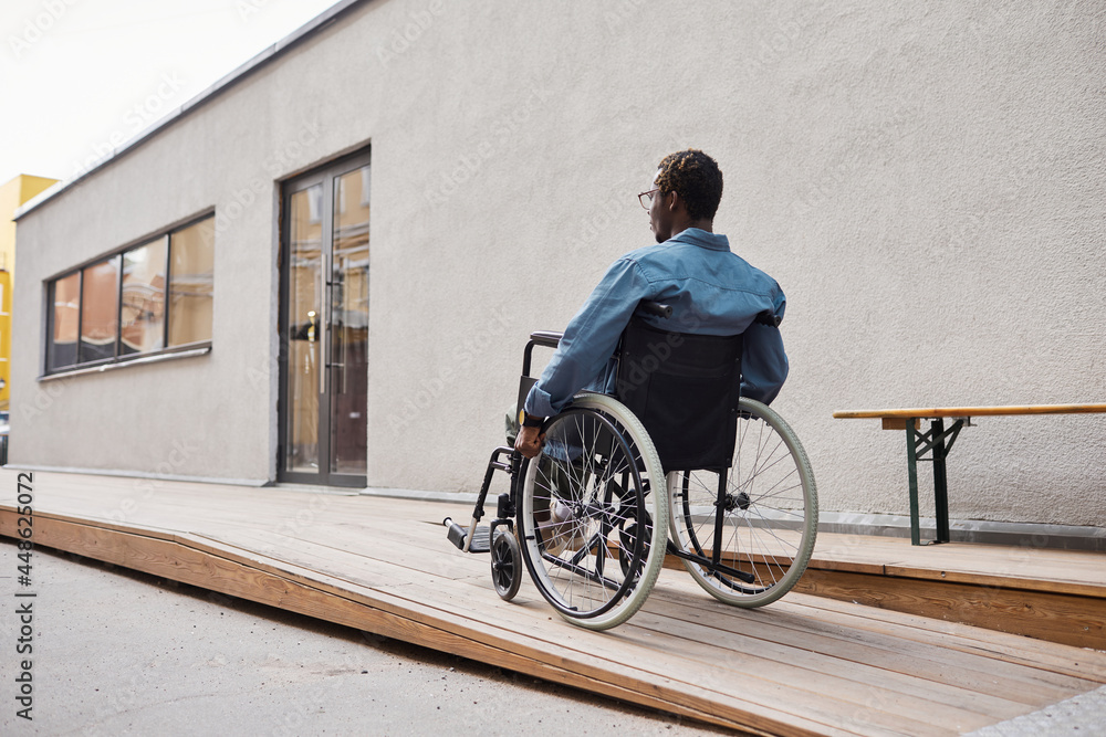 Rear view of handicapped African-American man in casual shirt using wheelchair ramp built by his house entrance Rear view of handicapped African-American man in casual shirt using wheelchair ramp built by his house entrance