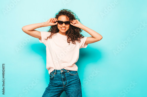 Beautiful black woman with afro curls hairstyle.Smiling model in white trendy t-shirt and jeans clothes. Sexy carefree female posing near blue in studio. Tanned and cheerful. In sunglasses