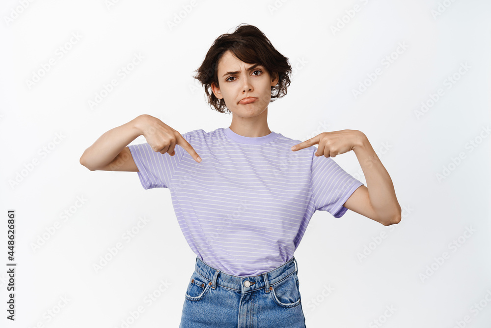Skeptical brunette girl pointing fingers at product, showing logo sale banner with upset, disappointed face, frowning and sulking unsure, standing over white background