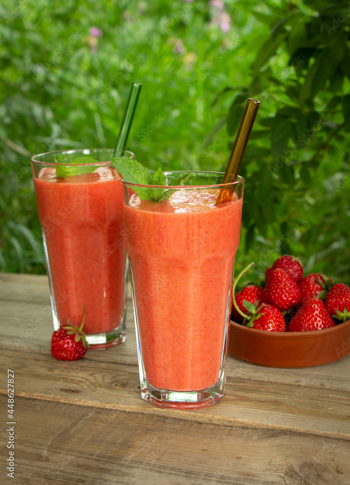 Two glasses with strawberry smoothie with mint leaves and glass straws on a wooden table in the garden Two glasses with strawberry smoothie with mint leaves and glass straws on a wooden table in the garden