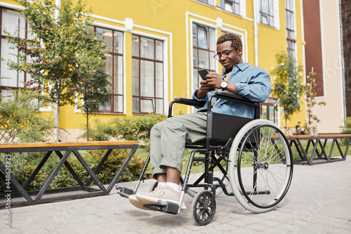Content handsome young black man with disability sitting in wheelchair and browsing Internet on smartphone during stroll