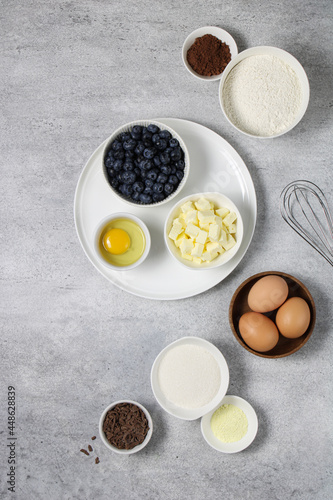 Cake ingredients  flour  butter  baking powder  chocolate  blueberries  eggs  cocoa  sugar  whisk on a light background. Ingredients for a chocolate blueberry pie recipe top view. 