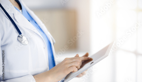 Unknown woman-doctor standing in sunny clinic and using tablet pc, close-up. Data and best service in medicine