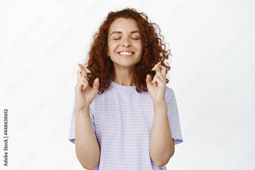 Fototapeta premium Hopeful young woman with red curly hair making wish, close eyes and cross fingers for good luck, wishing, anticipating positive results, white background
