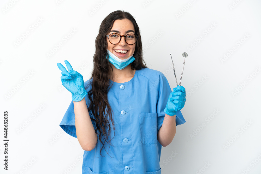 Young woman dentist holding tools isolated on white background smiling and showing victory sign Young woman dentist holding tools isolated on white background smiling and showing victory sign