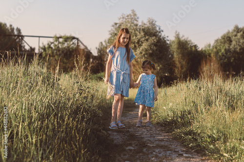 Two little girls walking in green meadow with wild grass. Sisters enjoy summer sunny day and beautiful nature together.