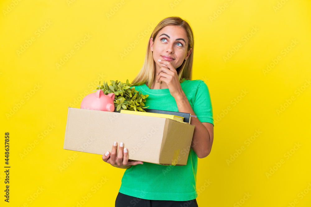Young Uruguayan girl making a move while picking up a box full of things isolated on yellow background looking up while smiling Young Uruguayan girl making a move while picking up a box full of things isolated on yellow background looking up while smiling