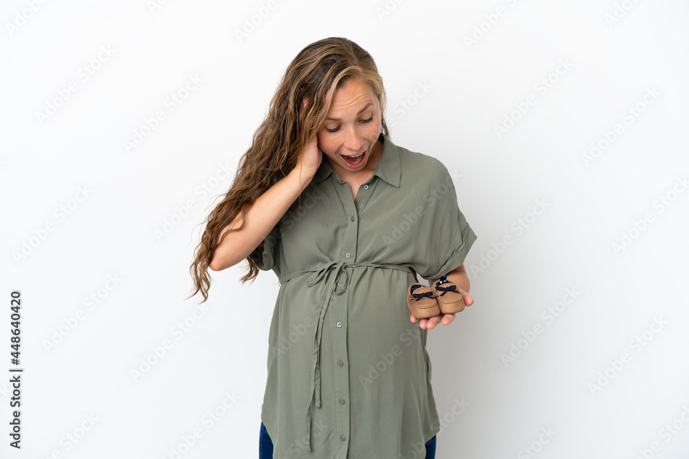 Young caucasian woman isolated on white background pregnant and holding baby booties with surprised expression