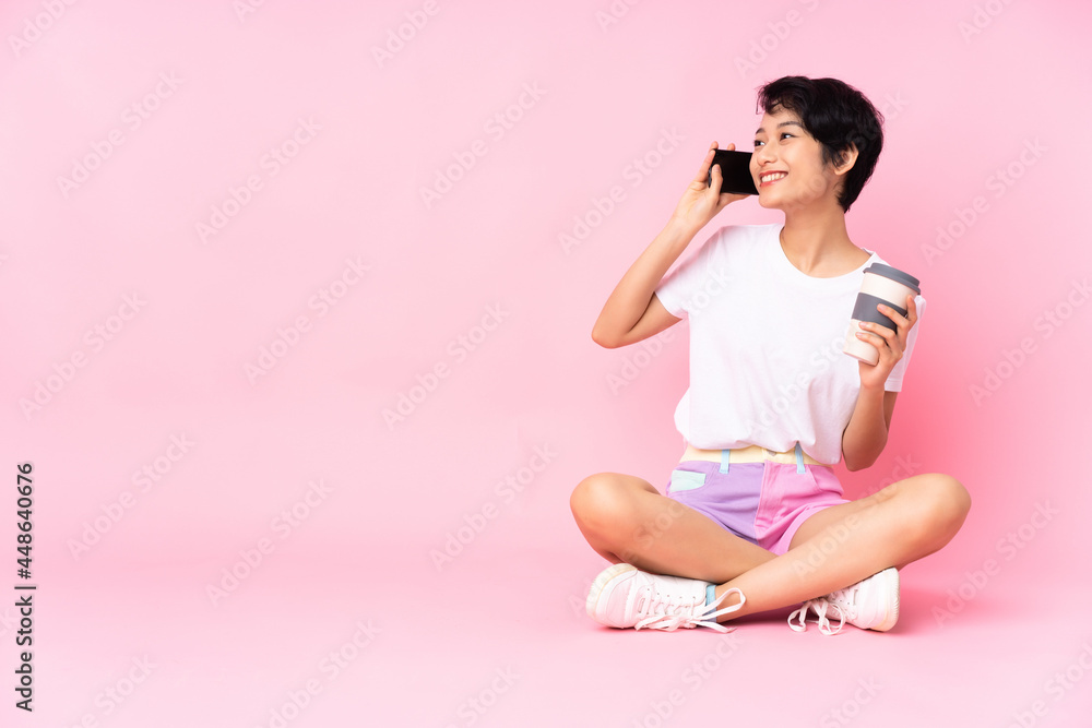 Young Vietnamese woman with short hair sitting on the floor over isolated pink background holding coffee to take away and a mobile Young Vietnamese woman with short hair sitting on the floor over isolated pink background holding coffee to take away and a mobile