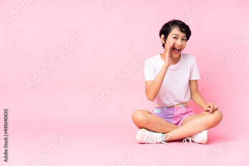 Young Vietnamese woman with short hair sitting on the floor over isolated pink background shouting with mouth wide open