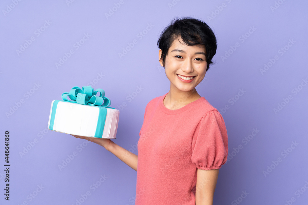 Young Vietnamese woman with short hair holding a big cake over isolated purple background smiling a lot Young Vietnamese woman with short hair holding a big cake over isolated purple background smiling a lot