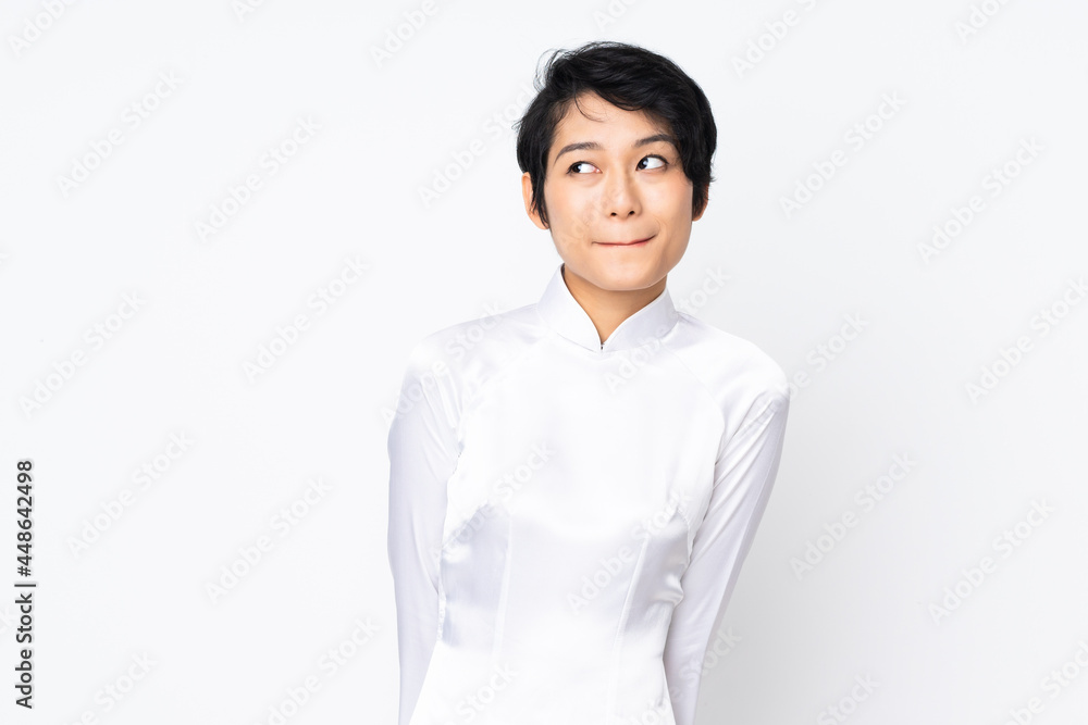 Young Vietnamese woman with short hair wearing a traditional dress over isolated white background having doubts while looking up Young Vietnamese woman with short hair wearing a traditional dress over isolated white background having doubts while looking up