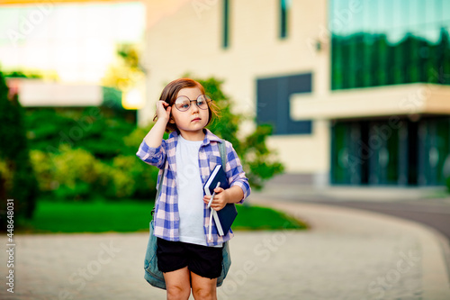 a little girl, a schoolgirl, wearing glasses, is standing near the school, with a backpack, holding a diary in her hands