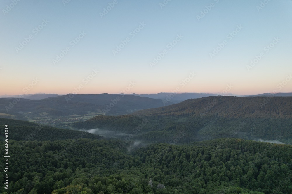 Summer panorama of Ukrainian mountain range. Beautiful summer scenery. Morning fog spreads on the mountain valley. Panoramic summer scene of Carpathian village. Amazing morning landscape of mountain. 