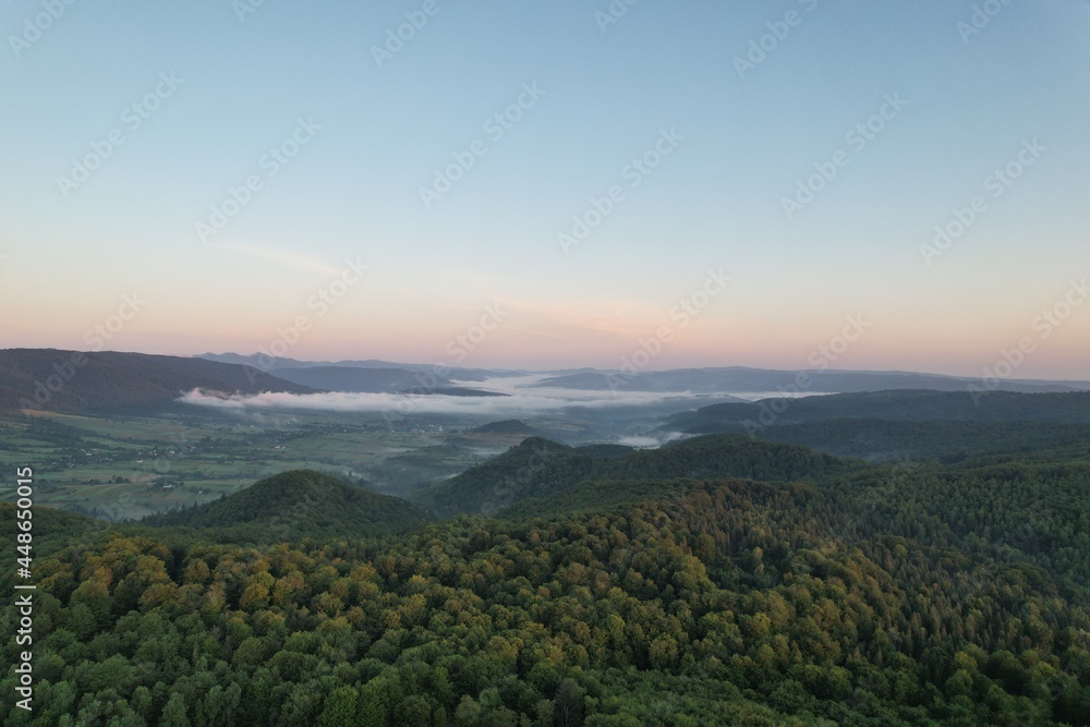 Summer panorama of Ukrainian mountain range. Beautiful summer scenery. Morning fog spreads on the mountain valley. Panoramic summer scene of Carpathian village. Amazing morning landscape of mountain. 