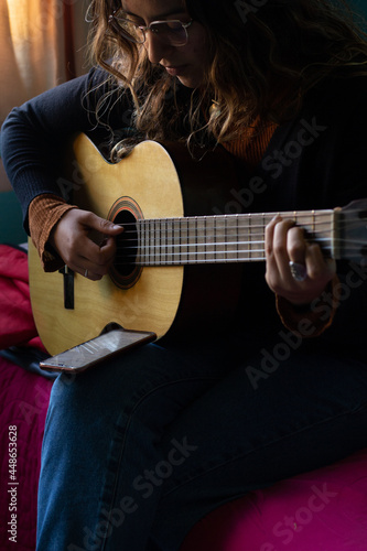 young woman playing guitar on her bedroom. She Is reading music of her phone. © Ruben