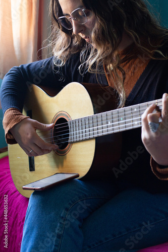 young woman playing guitar on her bedroom. She Is reading music of her phone. © Ruben