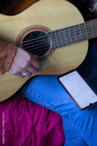 young hands of  woman playing guitar on her bedroom. She Is reading music of her phone. © Ruben