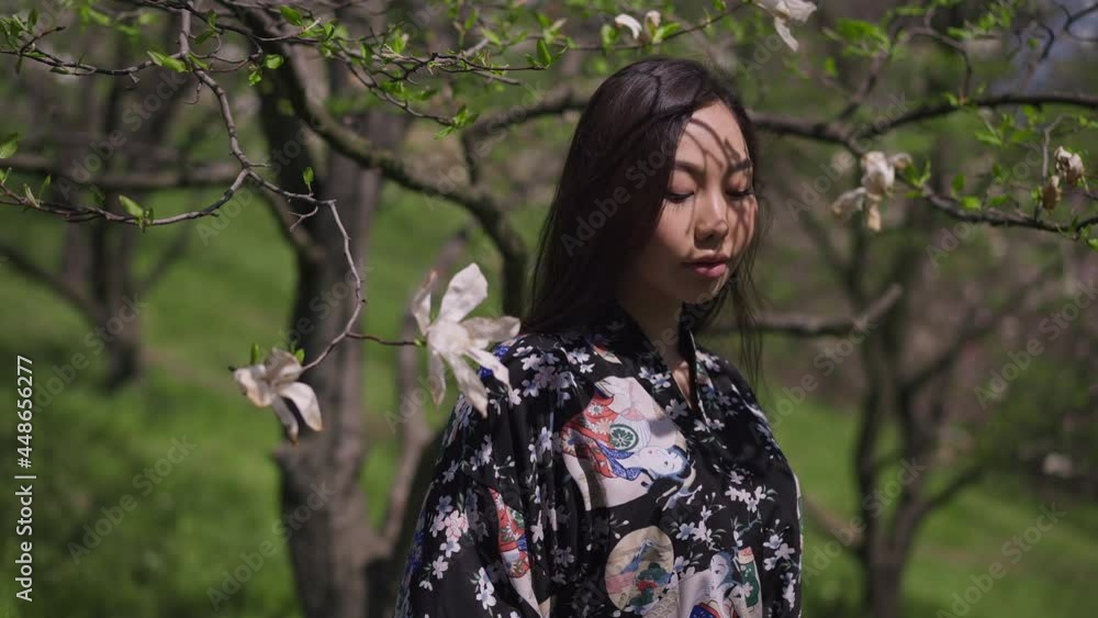Live camera zoom in to gorgeous Asian confident woman standing in sunlight in spring park with blooming trees. Portrait of relaxed happy young lady posing on sunny summer day outdoors. Slow motion Live camera zoom in to gorgeous Asian confident woman standing in sunlight in spring park with blooming trees. Portrait of relaxed happy young lady posing on sunny summer day outdoors. Slow motion