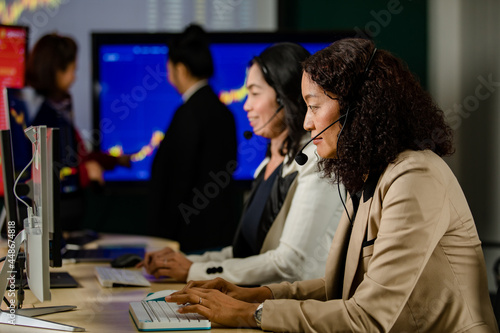 Asian female professional stock exchange manager sit on desk smile in front chart monitor look at camera while customer service operator female team working in blurred foreground