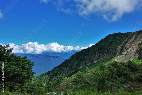The mountain slopes are covered with green vegetation. Picturesque cumulus clouds can be seen above the peaks of the ridge. Blue sky. A sunny summer day. Caucasus. Krasnaya Polyana