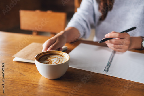 Closeup image of a woman drinking coffee and writing on a blank notebook on the table