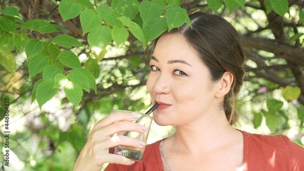 Girl sipping ice coffee through a straw of a glass with mean and sneaky eyes and naughty smile. Flirting behaviour of asian girl with white skin. Idyllic picnic scene portrait in a beautiful garden. Girl sipping ice coffee through a straw of a glass with mean and sneaky eyes and naughty smile. Flirting behaviour of asian girl with white skin. Idyllic picnic scene portrait in a beautiful garden.
