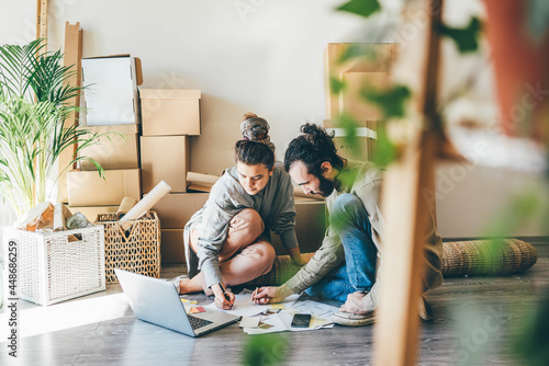 cheerful young family of man and woman wearing casual clothes makes project of decoration for new apartment on laptop sitting on floor near pile of boxes