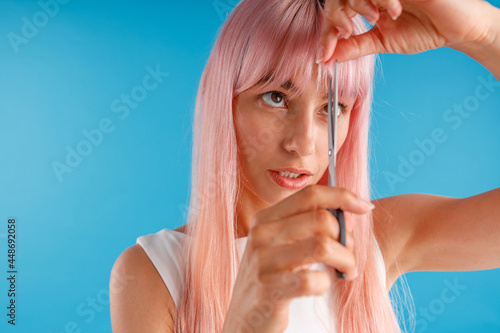 Portrait of cute young woman with pink hair cutting fringe herself using scissors, standing isolated over blue studio background. Beauty and hair care concept photo