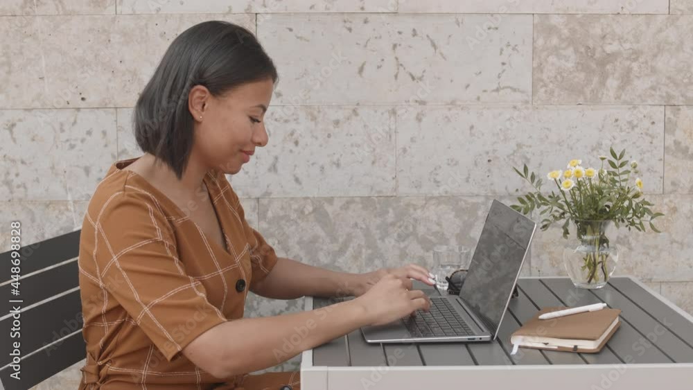 Side view medium of modern Mixed-Race businesswoman sitting at table outdoors in foreground of stone wall, typing on laptop computer, smiling Side view medium of modern Mixed-Race businesswoman sitting at table outdoors in foreground of stone wall, typing on laptop computer, smiling