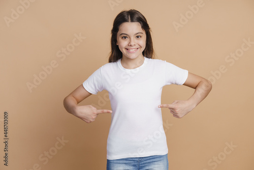 Positive, smiling teenage girl points to her white T-shirt, a month for advertising. Little girl on a brown background