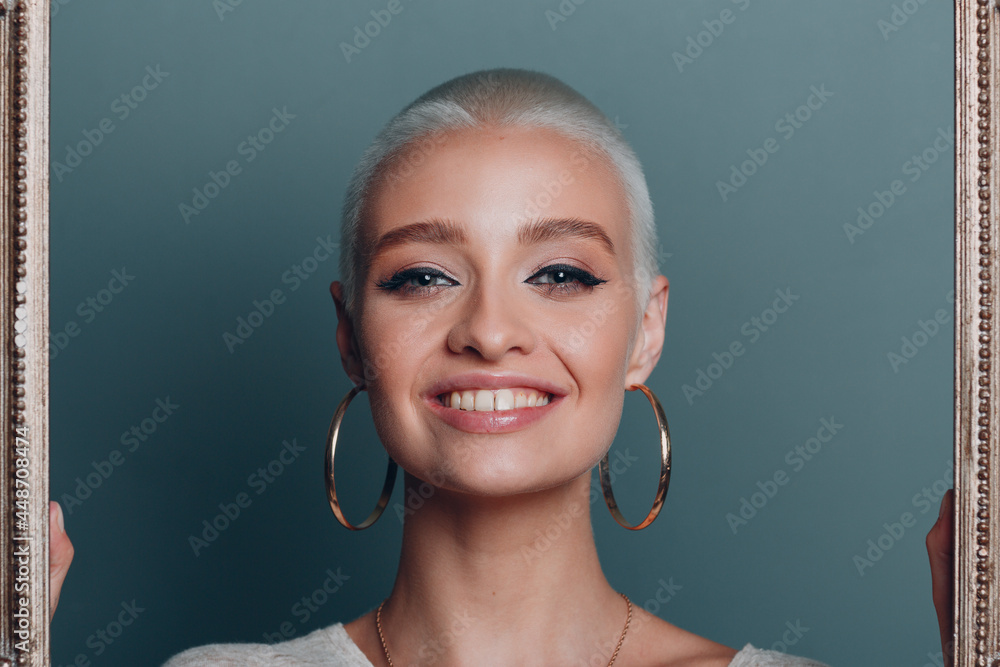 Millenial young woman with short blonde hair holds gilded picture frame in hands behind her face portrait Millenial young woman with short blonde hair holds gilded picture frame in hands behind her face portrait