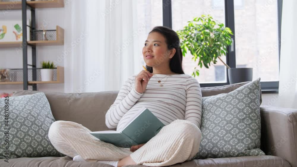 pregnancy, people and leisure concept - happy smiling pregnant asian woman sitting on sofa and writing to diary or notebook at home pregnancy, people and leisure concept - happy smiling pregnant asian woman sitting on sofa and writing to diary or notebook at home