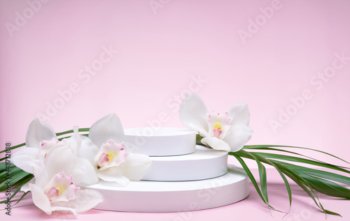 White geometric shapes podium for product display on pink background with orchid flowers and palm leaves. Monochrome stage  stand for product promotion in minimal style.
