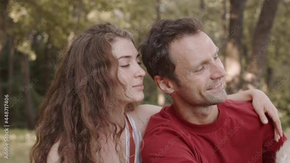 Medium close-up of cheerful Caucasian woman and man cuddling, sitting in summer park, petting cute Golden Retriever dog, talking and smiling Medium close-up of cheerful Caucasian woman and man cuddling, sitting in summer park, petting cute Golden Retriever dog, talking and smiling