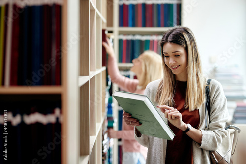 Smiling female student reading book cover while learning at university library.