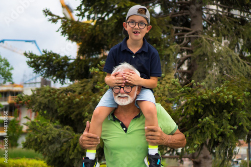 Grandpa is enjoying a walk in the park with his grandson, carrying him on his shoulders.