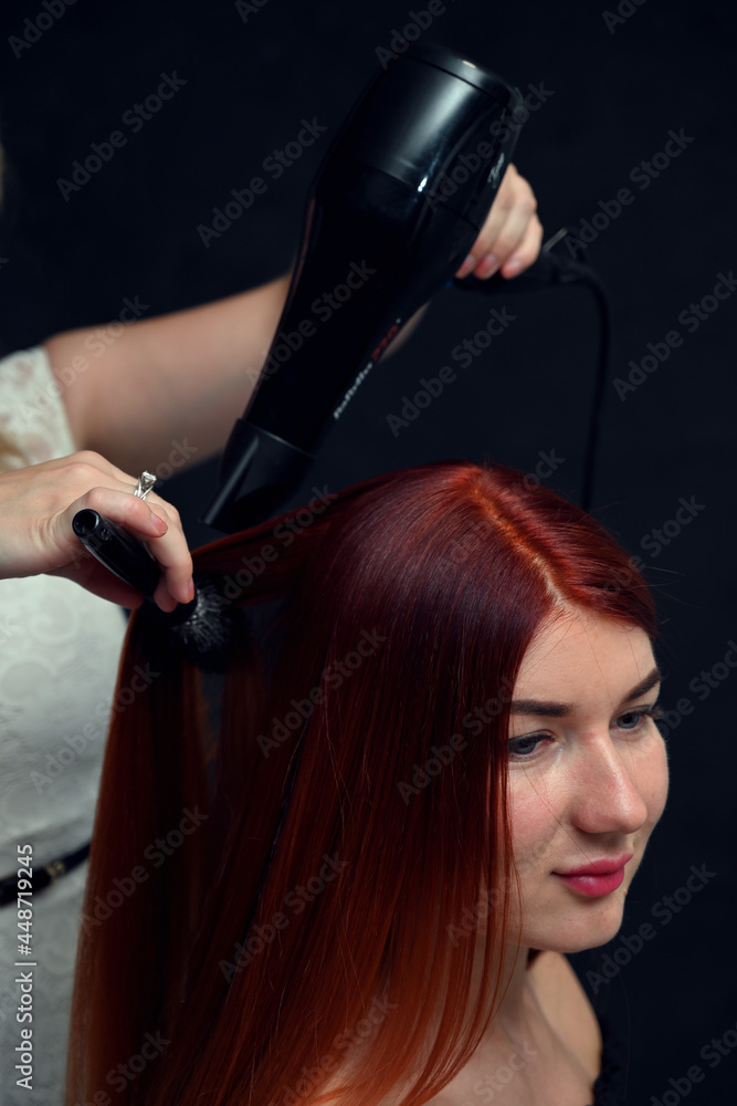Hairdresser working with red-haired model with hairdryer on gray background Hairdresser working with red-haired model with hairdryer on gray background