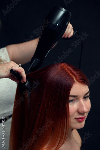 Hairdresser working with red-haired model with hairdryer on gray background